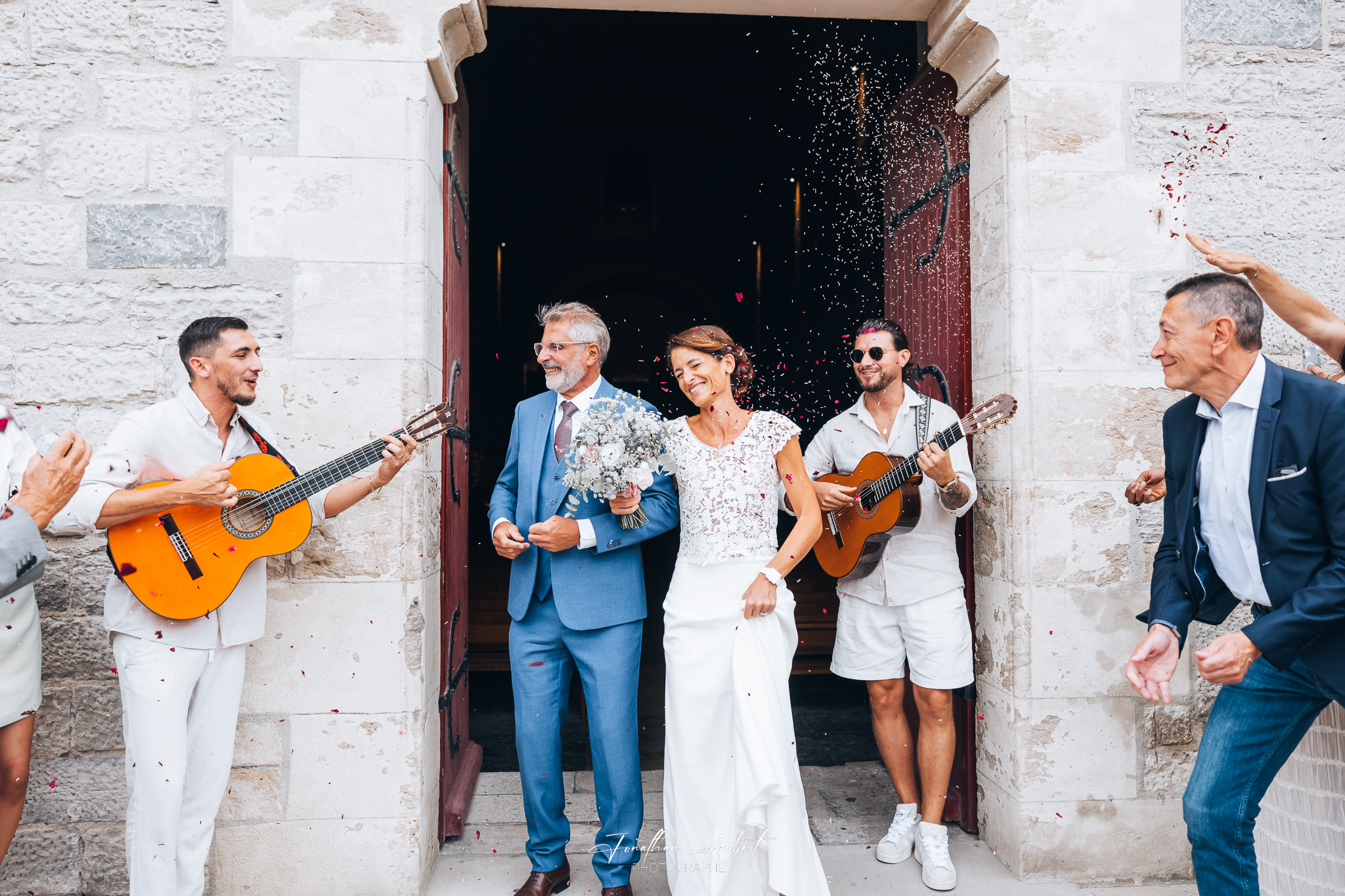 Sortie de mariage joyeuse avec les mariés entourés de musiciens jouant de la guitare, confettis en l'air, porte en pierre en arrière-plan