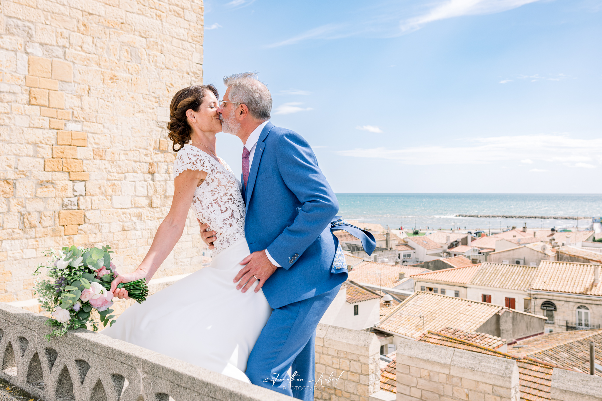 Couple de mariés s'embrassant sur un balcon en pierre avec vue sur la mer et les toits du village côtier