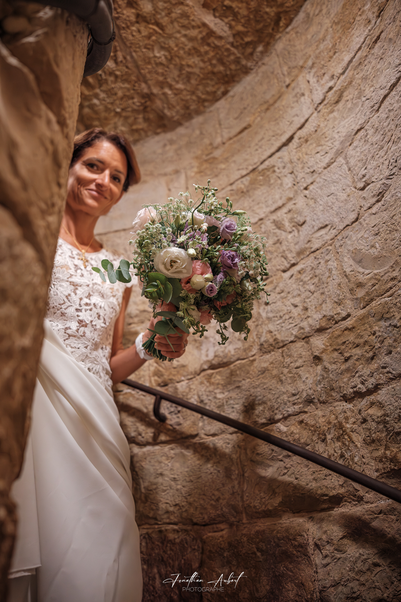 Mariée en robe blanche dentelle tenant un bouquet champêtre, posant dans une ruelle en pierre ancienne