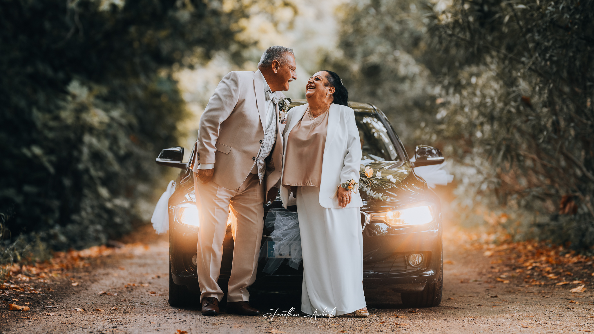Couple de mariés âgés riant ensemble devant une voiture, éclairage doré d'automne, allée bordée d'arbres