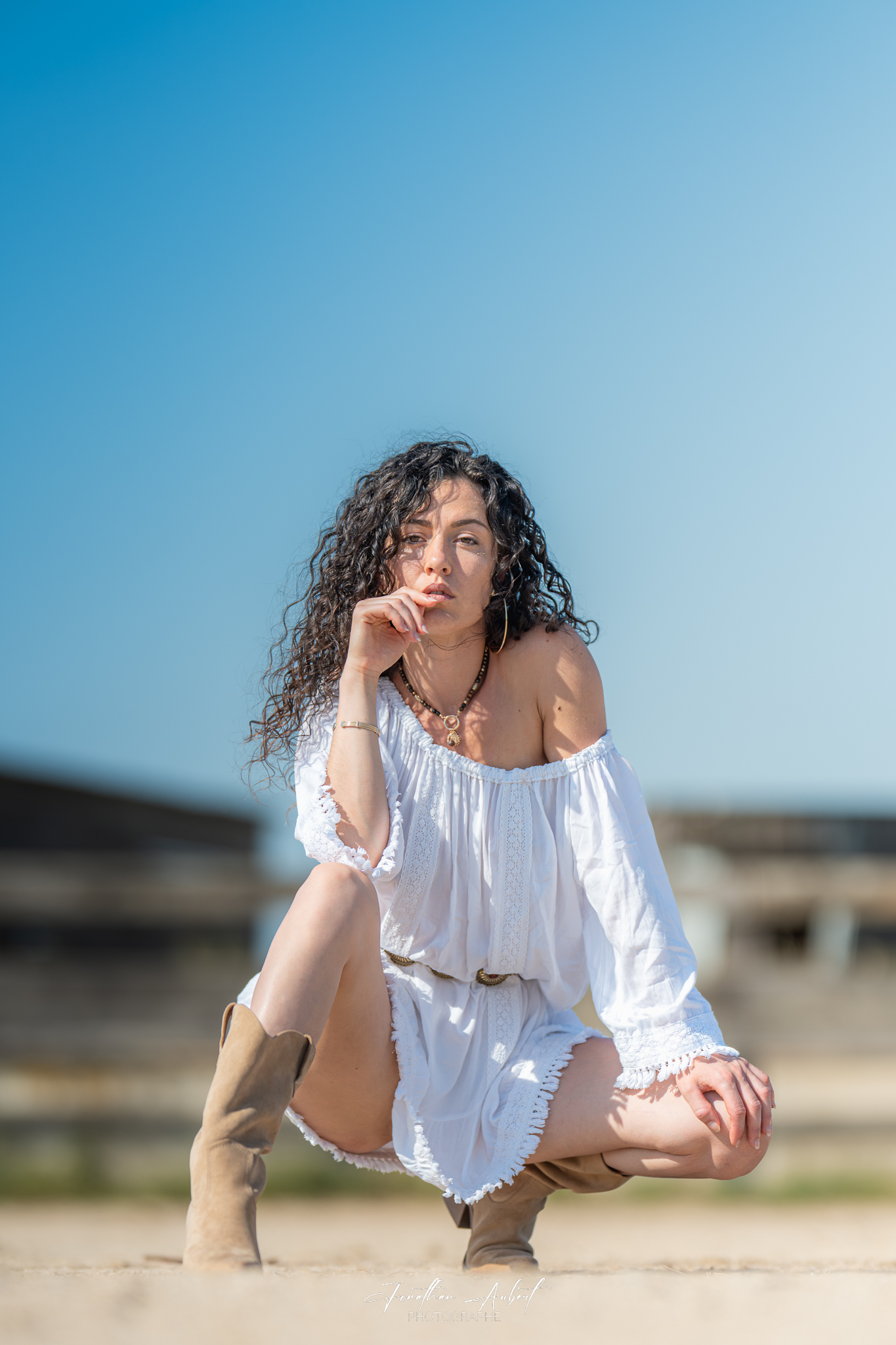 Portrait couleur d'une jeune femme aux cheveux bouclés en robe blanche légère, posant accroupie sous un ciel bleu clair